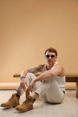 Moda modeli. Şapkalı ve güneş gözlüklü yakışıklı adam, bankın yanında bej arka planda poz veriyor, mesaj için yer var.