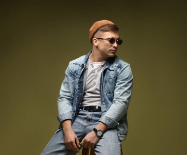 Moda modeli. Şapkalı, güneş gözlüklü ve ceketli yakışıklı adam koyu yeşil renkli arka plana karşı dışkı üzerinde poz veriyor.