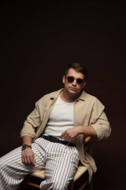 Moda modeli. Güneş gözlüklü yakışıklı siyah arka planda sandalyede poz veriyor.