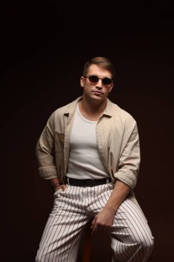 Moda modeli. Siyah arka planda taburede poz veren güneş gözlüklü yakışıklı bir adam.