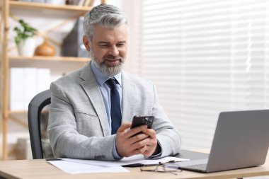 Ofisteki masada akıllı telefon kullanarak gülümseyen takım elbiseli işadamı.