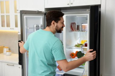 Mutfakta farklı gıda ürünleri olan açık buzdolabının yanındaki adam.