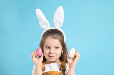 Paskalya kutlaması. Saçbantlı, tavşan kulaklı, açık mavi arka planda boyalı yumurtalar tutan gülümseyen küçük bir kız.