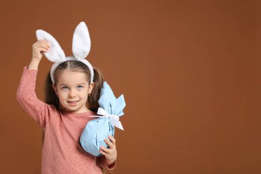 Paskalya kutlaması. Saçbantlı, tavşan kulaklı gülümseyen küçük bir kız kahverengi arka planda hediye tutuyor, mesaj için yer var.