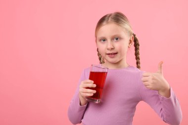 Elinde bir bardak nar suyu olan küçük bir kız pembe arka planda baş parmağını gösteriyor. Metin için boşluk