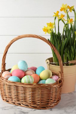 Hasır sepete boyanmış Paskalya yumurtaları ve beyaz mermer masa üzerindeki güzel nergisler, yakın plan.