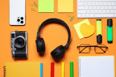 Kulaklıklar, bilgisayar klavyesi, akıllı telefon, klasik kamera, turuncu arka planda gözlük ve kırtasiye malzemesi.