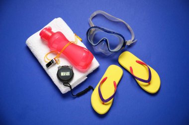 Dalış maskesi, terlikler, bir şişe su, havlu, dijital kronometre ve mavi arka planda ıslık. Yüzme aksesuarı