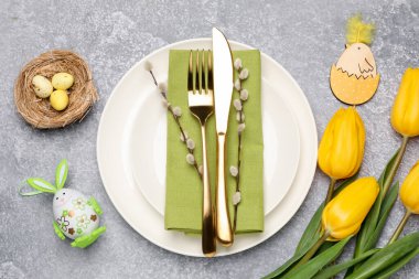 Paskalya kutlaması. Lale çiçekleri, çatal bıçak takımı, söğüt dalları ve gri desenli masaya boyanmış yumurtalarla süslü bir yer.