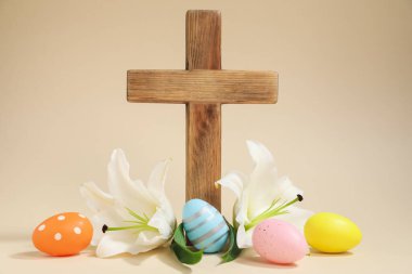 Paskalya. Ahşap haç, boyalı yumurta ve bej arka planda güzel zambak çiçekleri, yakın plan.