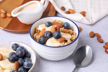 Yulaf gevreği, badem sütü, fındık, muz ve yaban mersini açık gri masada, yakın plan.