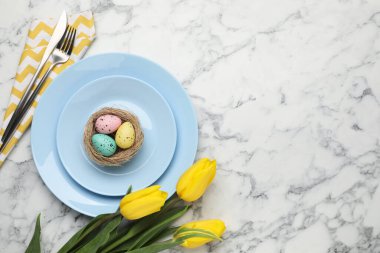 Paskalya kutlaması. Çatal bıçak takımı, çiçekler ve beyaz mermer masaya boyanmış yumurtalar, düz yatak. Metin için boşluk