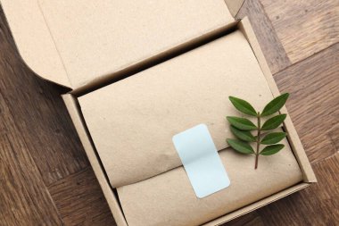 Ambalajlı karton kutu, boş çıkartma ve ahşap masa üzerinde yeşil yapraklar, üst görünüm
