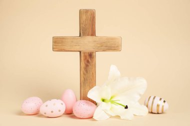 Paskalya. Ahşap haç, boyalı yumurta ve bej arka planda güzel zambak çiçeği, yakın plan.