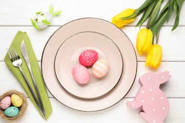 Paskalya kutlaması. Çatal bıçak, çiçek ve beyaz ahşap masaya boyanmış yumurtalarla şenlikli bir ortam.