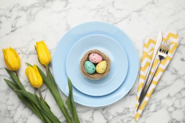 Paskalya kutlaması. Çatal bıçak takımı, çiçekler ve beyaz mermer masaya boyanmış yumurtalar, düzlük