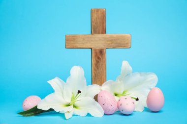 Paskalya. Ahşap haç, boyanmış yumurtalar ve açık mavi arka planda güzel zambak çiçekleri, yakın plan.