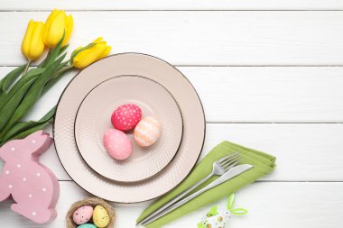 Paskalya kutlaması. Çatal bıçak takımı, çiçekler ve beyaz ahşap masaya boyanmış yumurtalar, düz yatak. Metin için boşluk