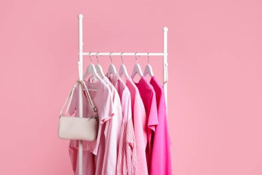 Farklı kıyafetler ve çantalar pembe arka planda asılı, yakın plan.