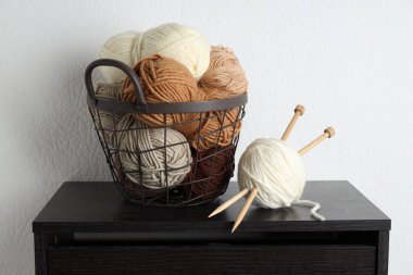 Beyaz duvarın yanındaki siyah ahşap şifoniyerde örgü iplikleri ve iğneler var.