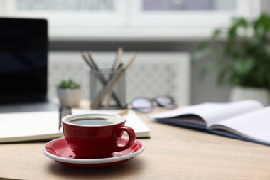 Ofisteki ahşap masada bir fincan kahve, dizüstü bilgisayar, bardak, kırtasiye ve çiçek dekoru, yakın plan. Metin için boşluk