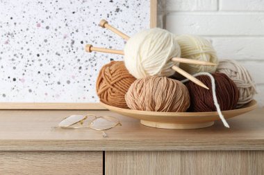 Örgü iplikleri, iğneler ve gözlükler beyaz tuğla duvarın yanındaki ahşap masada, yakın plan.