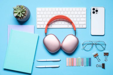Kulaklıklar, akıllı telefon, bilgisayar klavyesi, gözlükler, kırtasiye malzemeleri ve açık mavi arka planda çiçek dekoru, düz konum