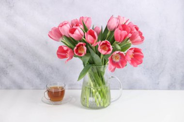 Vazoda pembe laleler ve gri desenli duvarın yanındaki beyaz masada bir fincan çay.