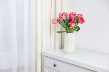 Beyaz duvarın yanındaki çekmece çekmecesinde vazoda duran pembe laleler. Metin için boşluk