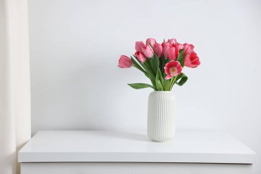 Beyaz duvarın yanındaki çekmecelerin üzerinde vazoda pembe laleler var.