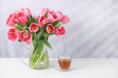 Vazoda pembe laleler ve gri desenli duvarın yanındaki beyaz masada bir fincan çay.