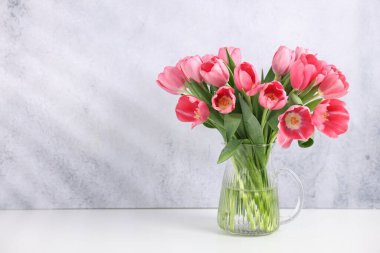 Bouquet of pink tulips in vase on white table near grey textured wall. Space for text