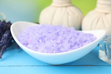 Kase içinde mor deniz tuzu, bitkisel torbalar ve lavanta çiçekleri açık mavi ahşap masa üzerinde bulanık yeşil arka plan, yakın çekim