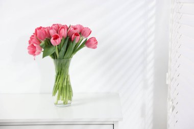 Beyaz duvarın yanındaki çekmece çekmecesinde vazoda duran pembe laleler. Metin için boşluk