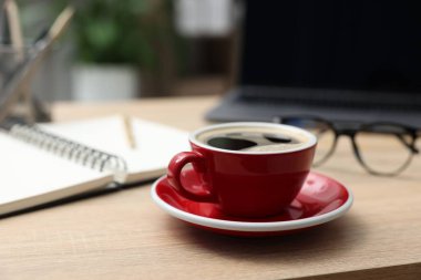 Ofisteki ahşap masaya bir fincan kahve, laptop, kırtasiye ve bardaklar.