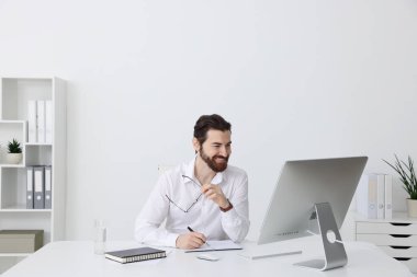 İşadamı bilgisayarla çalışıyor ve beyaz duvarın yanındaki masada yazı yazıyor.