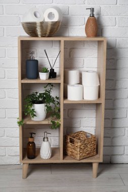 Tuvalet kağıdı ruloları, sabun, aroma yayıcı ve tuğla duvarın yanındaki raflarda bulunan ev bitkisi.