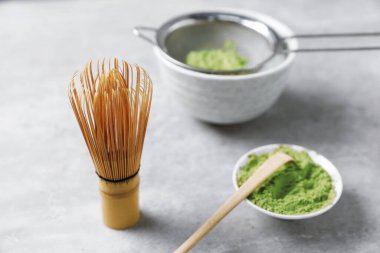 Green matcha powder in bowl, scoop, bamboo whisk and sifter on gray textured table, closeup