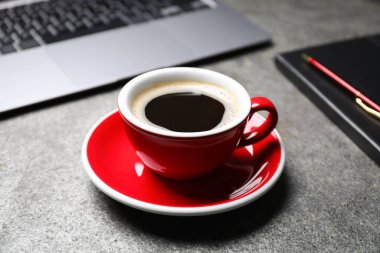 Gri masada bir fincan aromatik kahve, dizüstü bilgisayar ve ofis malzemeleri, yakın plan.