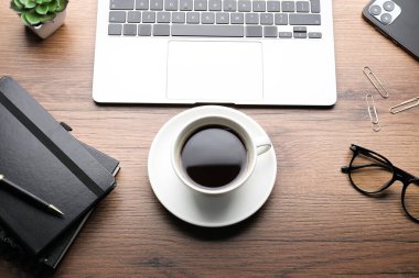 Bir fincan aromatik kahve, dizüstü bilgisayar ve ofis gereçleri ahşap masada, düz yerde.