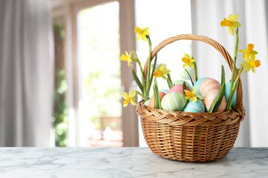 Hasır sepetin içinde paskalya yumurtaları ve nergisler bulanık pencereye karşı masada. Metin için boşluk