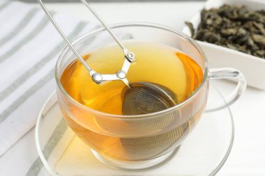 Bir fincan çay, kuru yapraklar ve beyaz ahşap masada metal aşılayıcı, yakın plan.