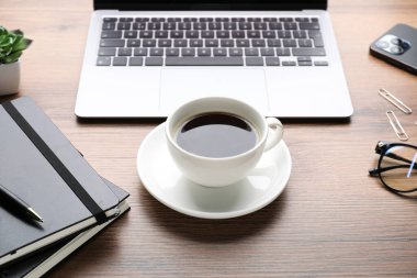 Bir fincan aromatik kahve, dizüstü bilgisayar ve ofis malzemeleri ahşap masada, yakın plan.