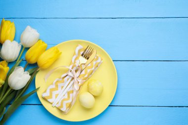 Paskalya kutlaması. Çatal bıçak takımları, laleler ve açık mavi ahşap masa üzerine boyanmış yumurtalar. Metin için boşluk