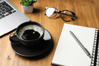 Bir fincan kahve, bardak, dizüstü bilgisayar, kırtasiye ve çiçek dekoru ahşap masada, yakın plan.