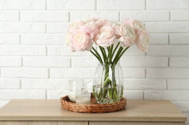 Güzel ranunculus çiçekleri cam vazoda ve mumlar ahşap çekmecede, beyaz tuğla duvarın yanında.
