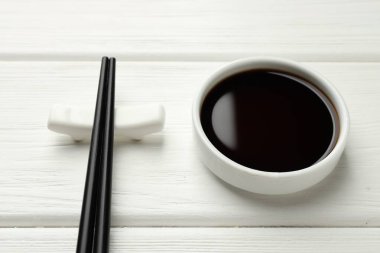 Soy sauce in bowl and chopsticks on white wooden table, closeup