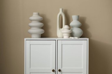 Bej duvarın yanındaki beyaz dolapta güzel vazolar var.