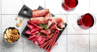 Şarküteri tahtası, farklı pişirilmiş et, sosis ve beyaz fayanslı masa üzerinde şarap.