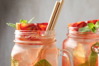 Tasty drink with Sicilian orange, ice cubes and mint in mason jars on blurred grey background, closeup